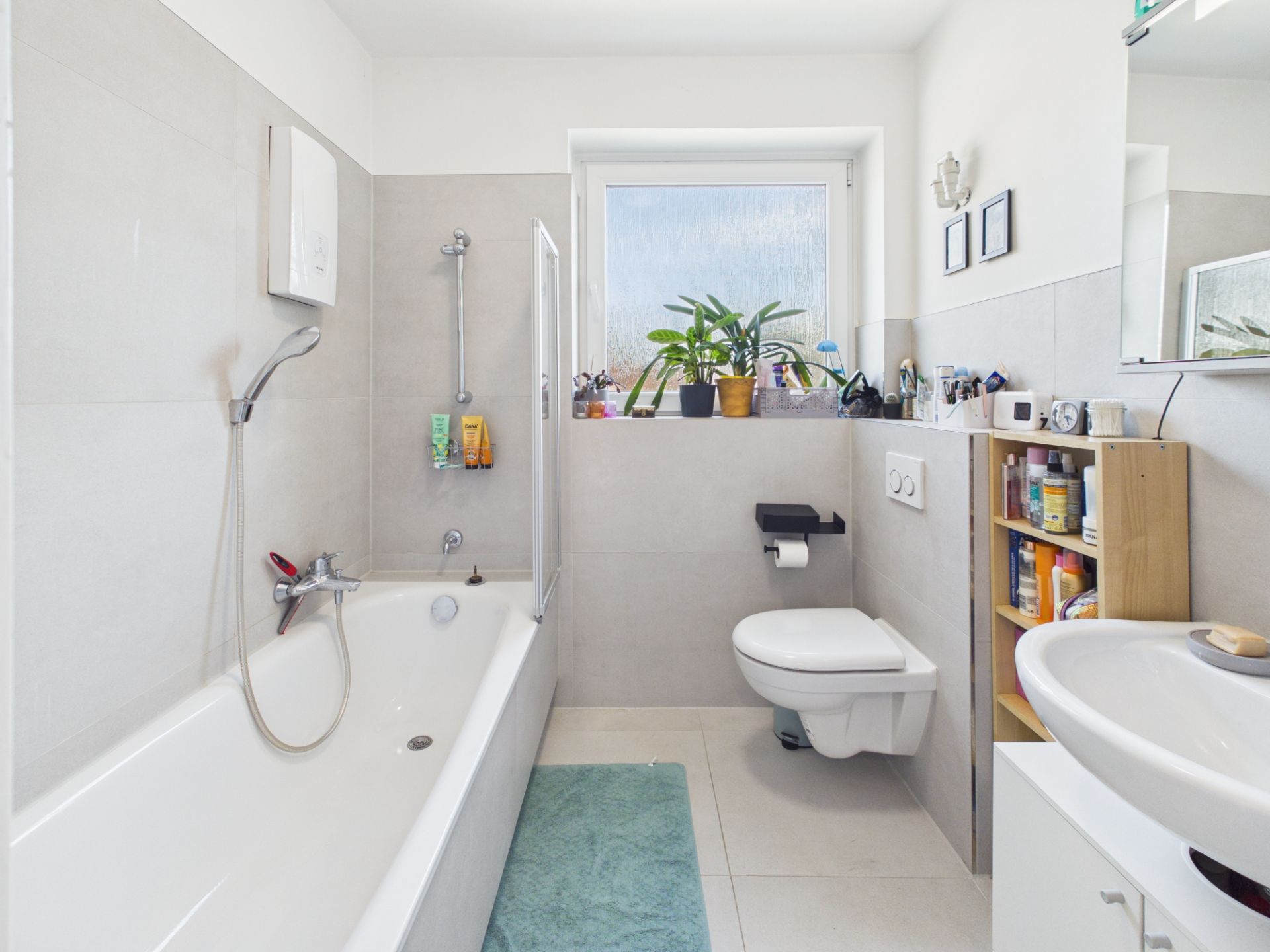 Helles Badezimmer mit Badewanne, Waschbecken, Toilette, Regal und Fenster mit Pflanzen.