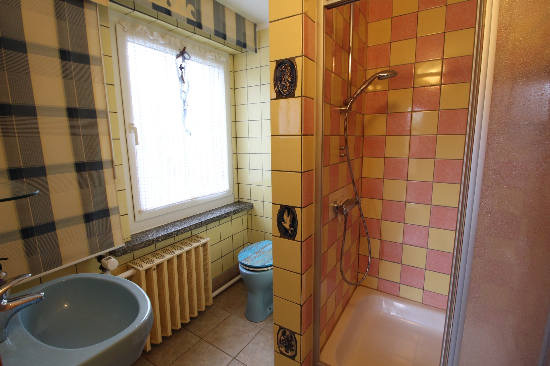 Kleines Badezimmer mit Dusche, blauem Waschbecken und Toilette, gelben und rosa Fliesen.