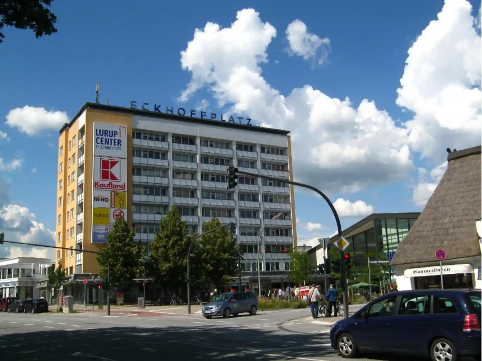 Mehrstöckiges Gebäude mit großen Fenstern, Geschäftsschildern und Bäumen an einer Straßenkreuzung.