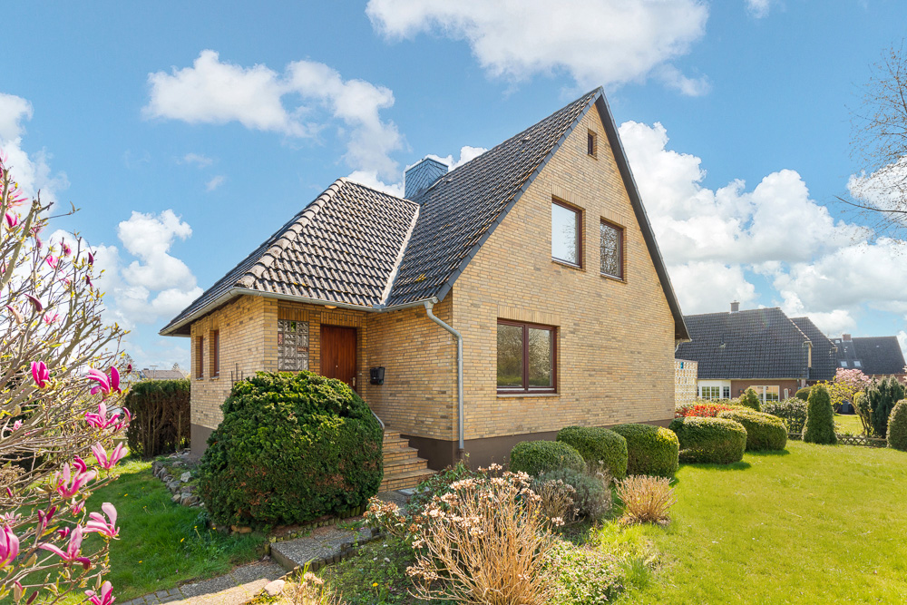 Einfamilienhaus aus gelben Ziegeln mit Garten und Sträuchern bei sonnigem Wetter.