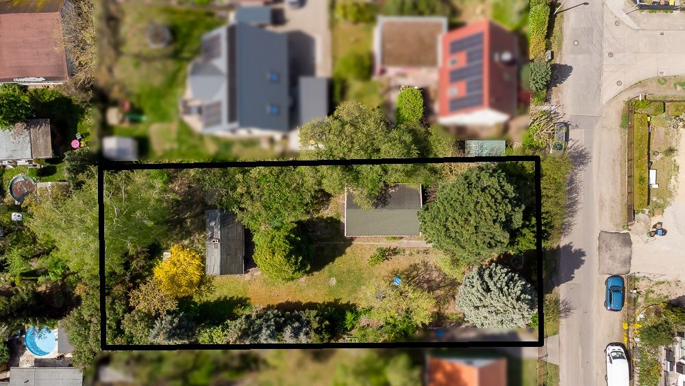 Luftaufnahme eines rechteckigen Grundstücks mit viel Grün und zwei Gebäuden, von Straßen und Nachbarhäusern umgeben.