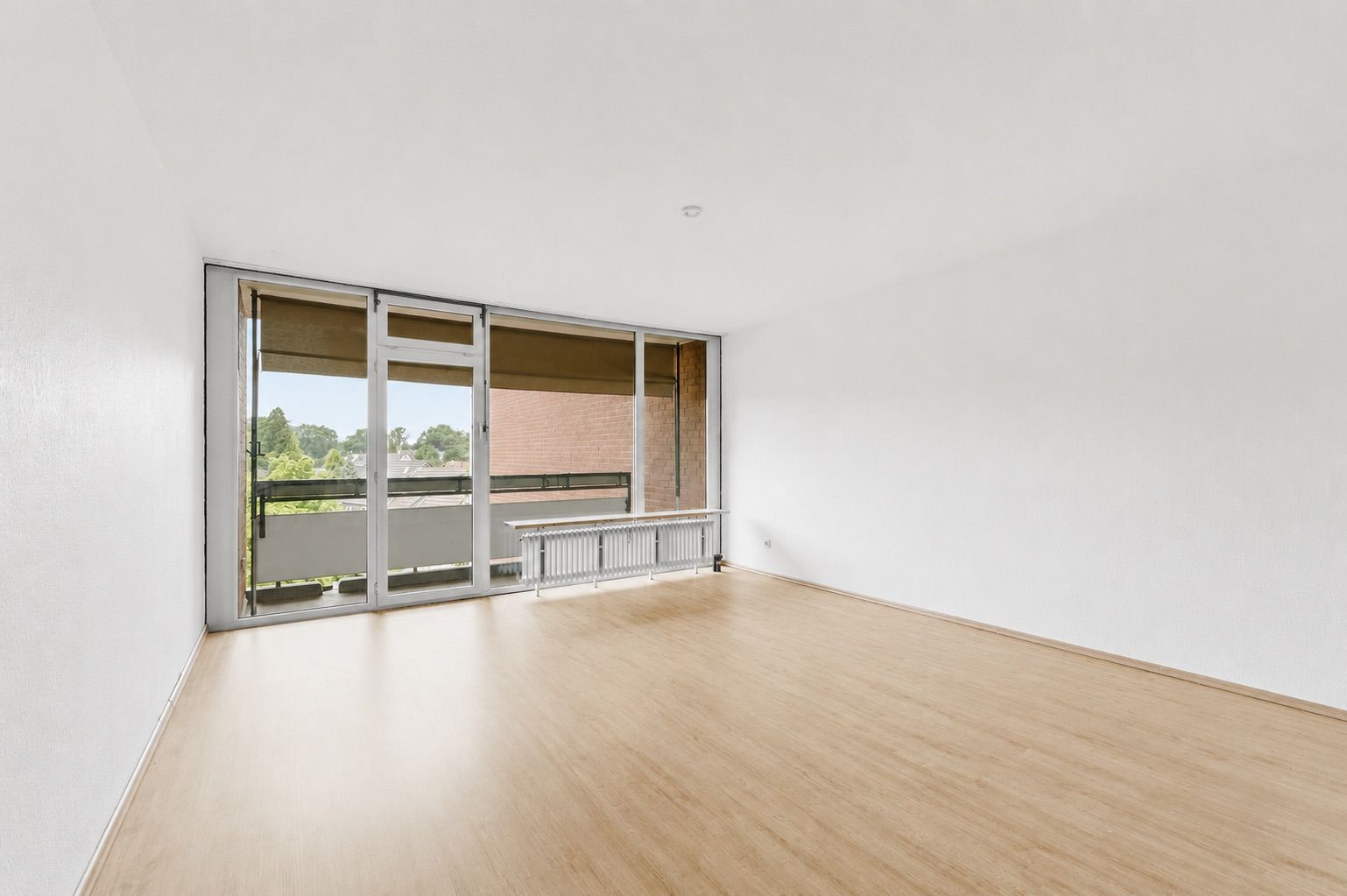 Leeres, helles Zimmer mit großem Fenster und Holzboden, Blick auf Balkon und grüne Bäume.