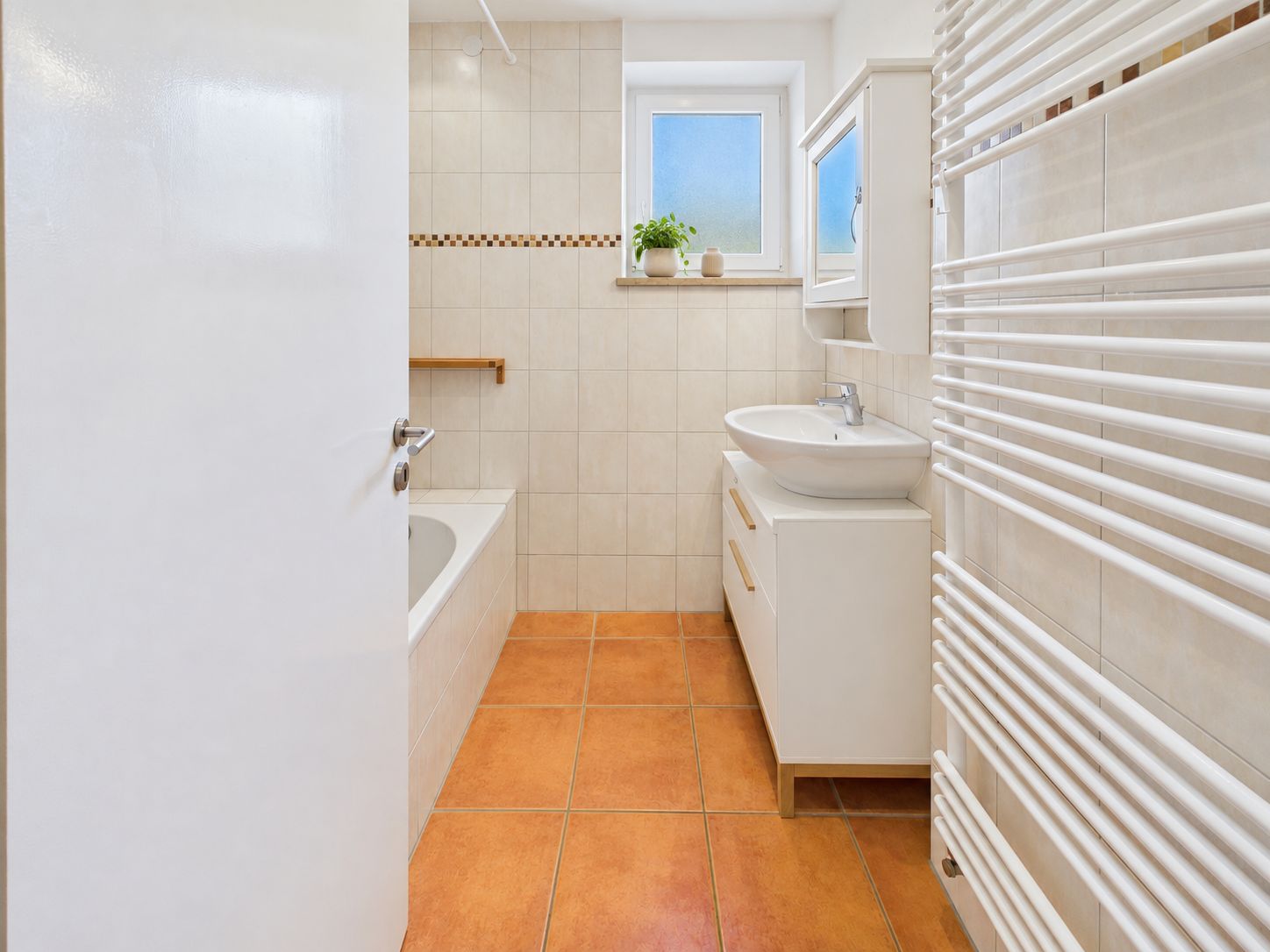 Modernes Badezimmer mit Badewanne, Waschbecken, Handtuchheizkörper und Fenster mit Pflanze.