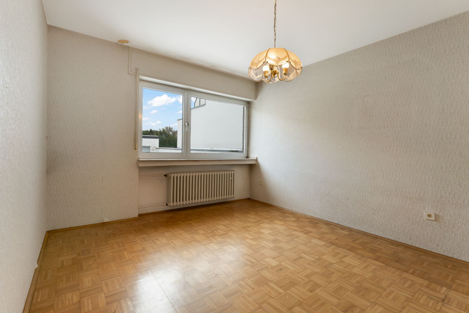 Leerer Raum mit Parkettboden, großem Fenster, Heizung und Deckenlampe.