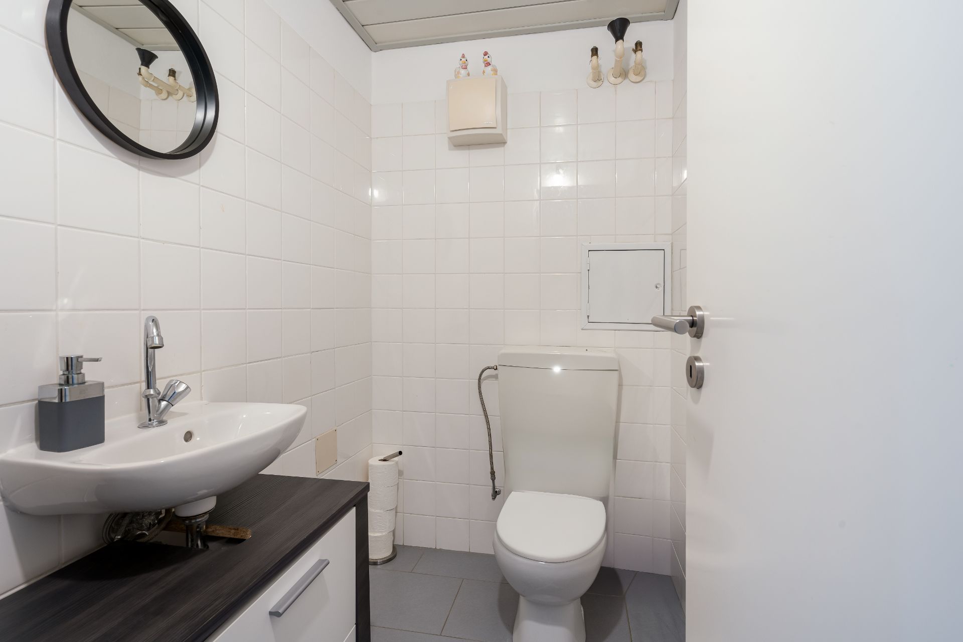 Kleines Badezimmer mit Toilette, Waschbecken, Spiegel und weiß gefliesten Wänden.