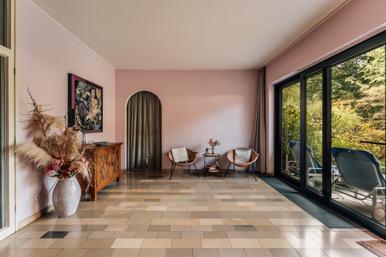 Heller Raum mit rosa Wänden, zwei Sesseln, großem Fenster und Zugang zur Terrasse mit Gartenblick.