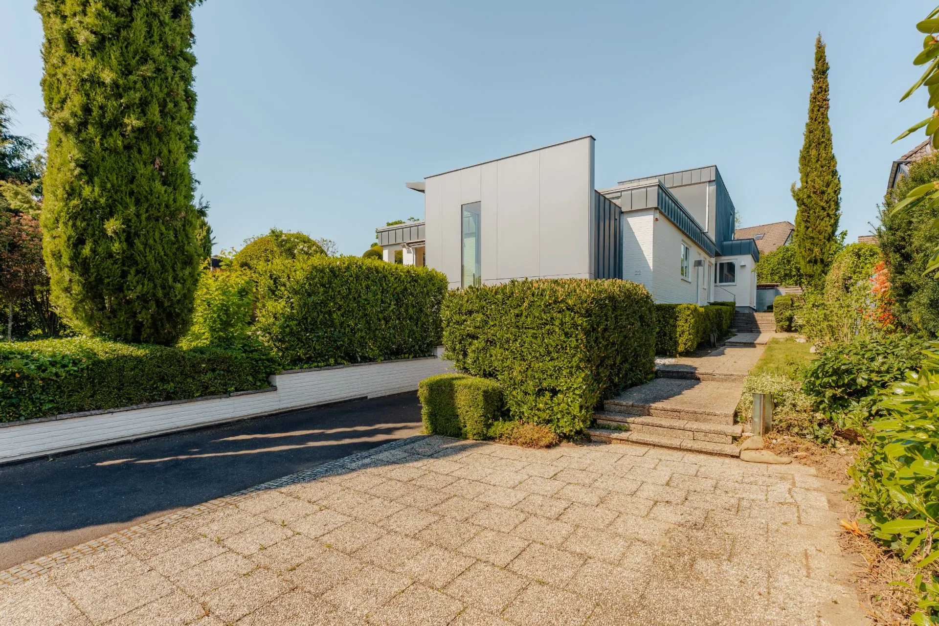 Modernes Wohnhaus mit grauer Fassade, Einfahrt und gepflegtem Garten, von Hecken umgeben.