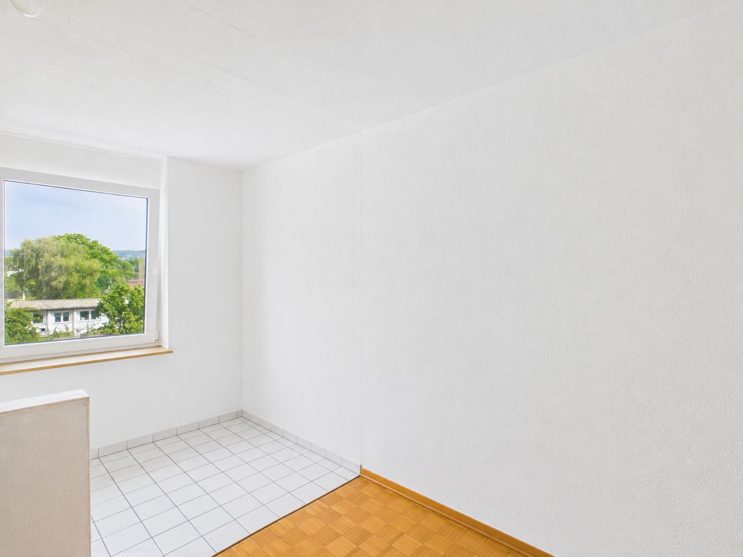 Heller Raum mit weißem Wänden, Parkett- und Fliesenboden, Fenster mit Blick ins Grüne.