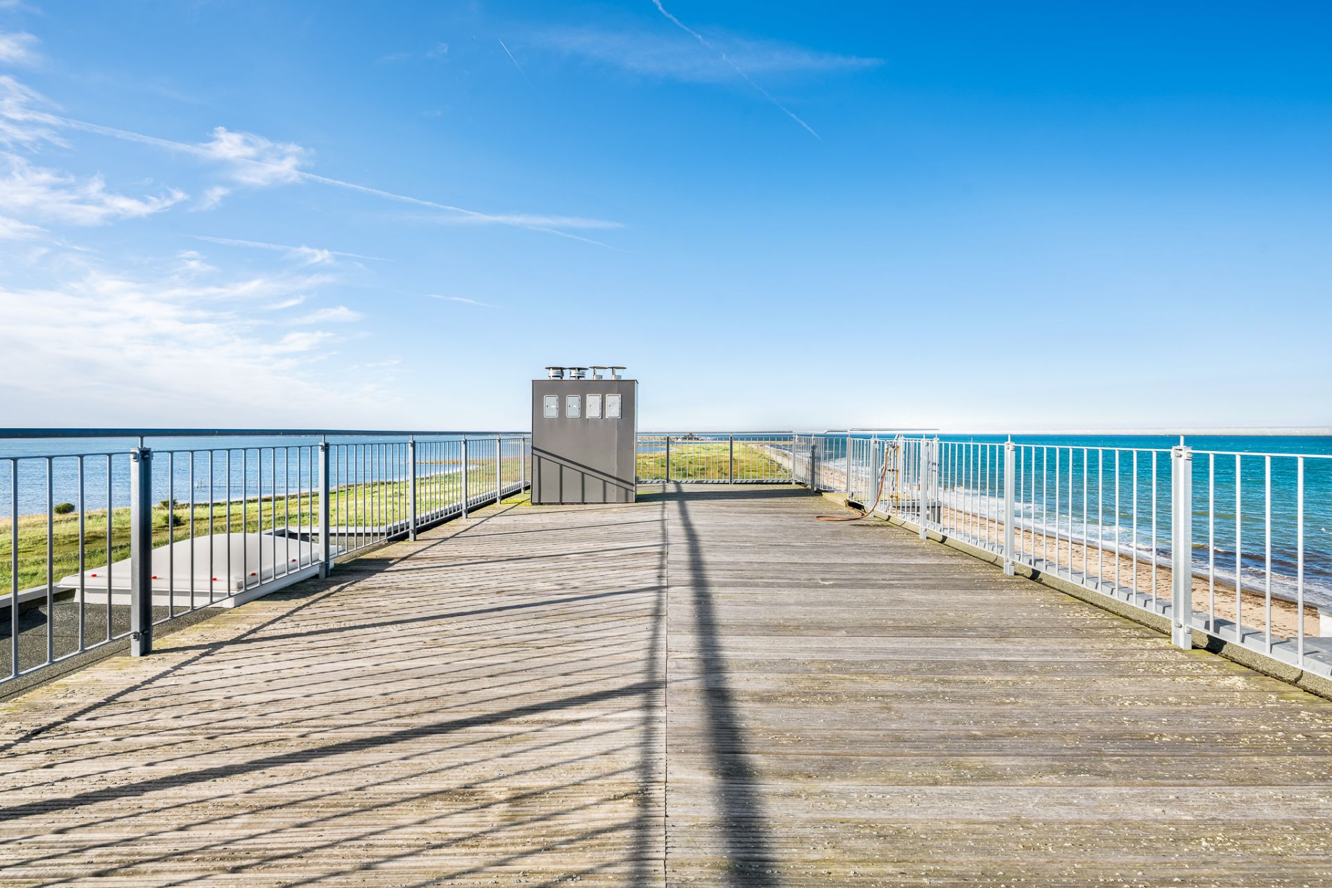 Große Holzterrasse mit Metallgeländer und Blick aufs Meer bei klarem Himmel.