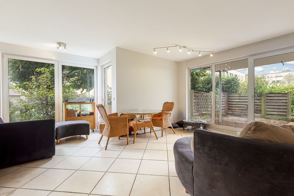 Helles Wohnzimmer mit großen Fenstern, zwei Sofas, Korbstühlen und Fliesenboden.