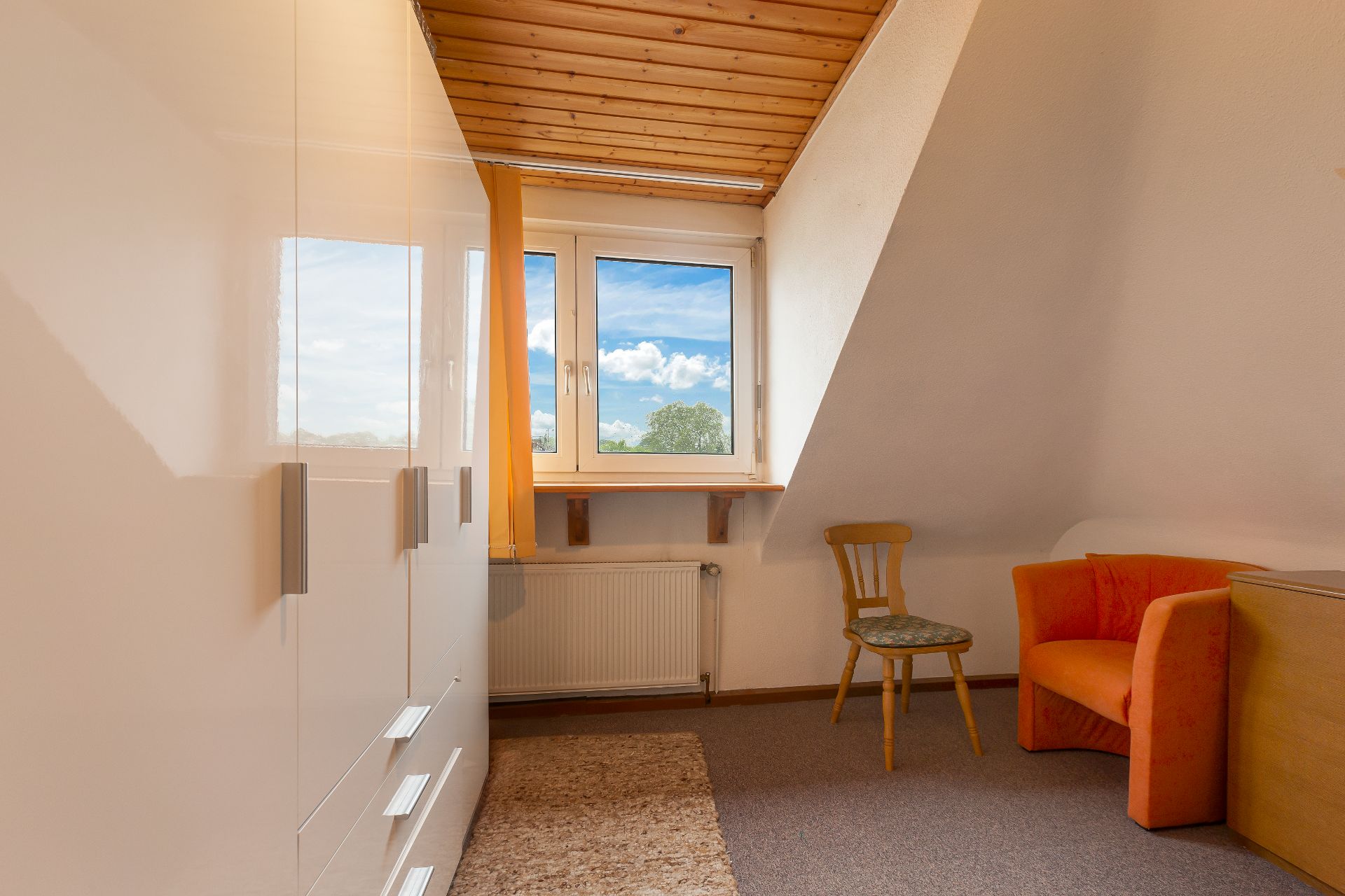 Kleines Zimmer mit weißem Schrank, Fenster, Holzstuhl und orangem Sessel unter Dachschräge.