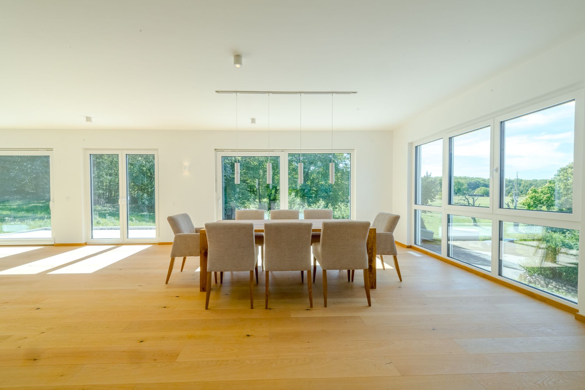 Heller Essbereich mit Holzboden, großem Tisch, acht Stühlen und großen Fenstern mit Blick ins Grüne.