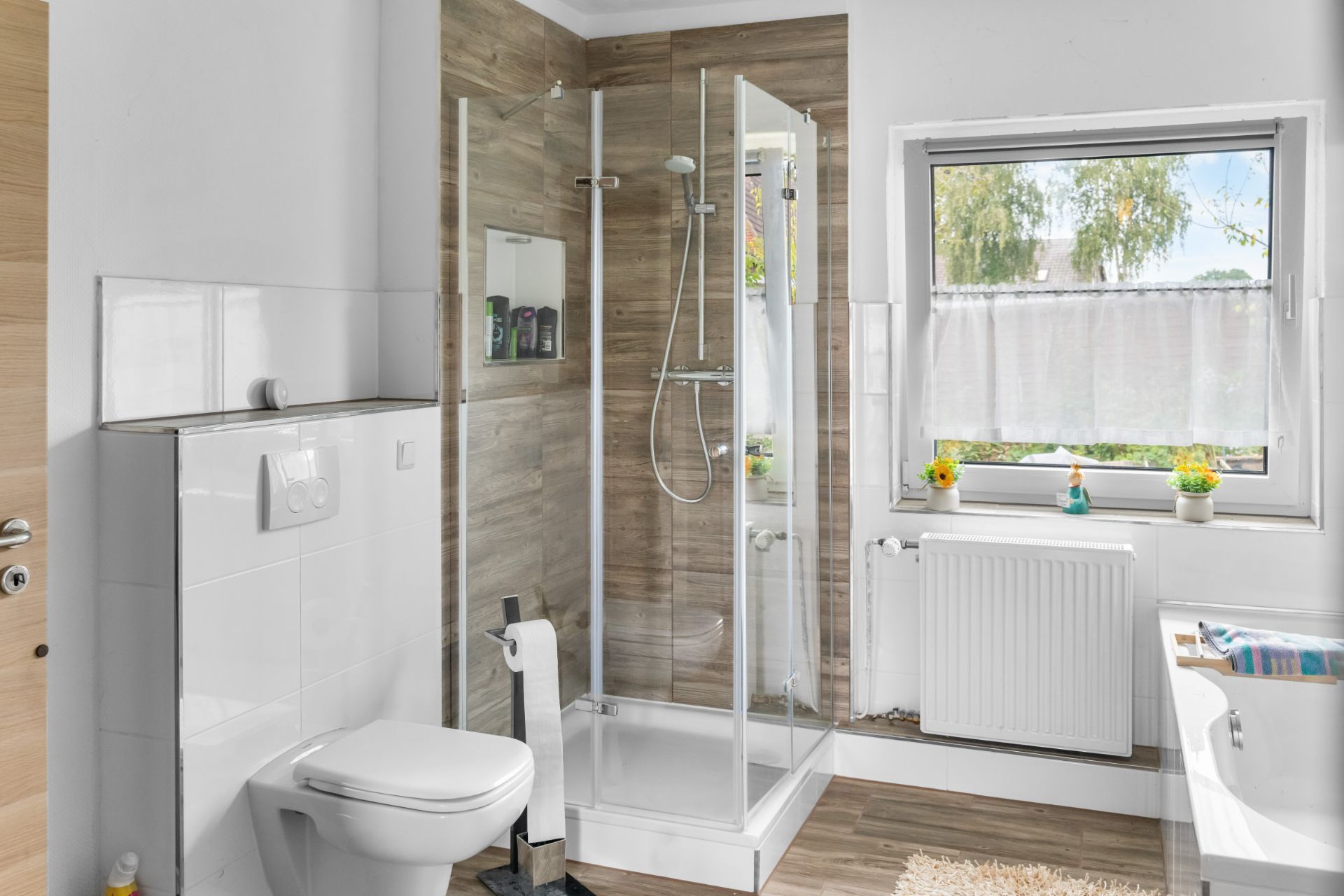 Modernes Badezimmer mit Dusche, Toilette, Fenster und Badewanne auf hellem Holzboden.