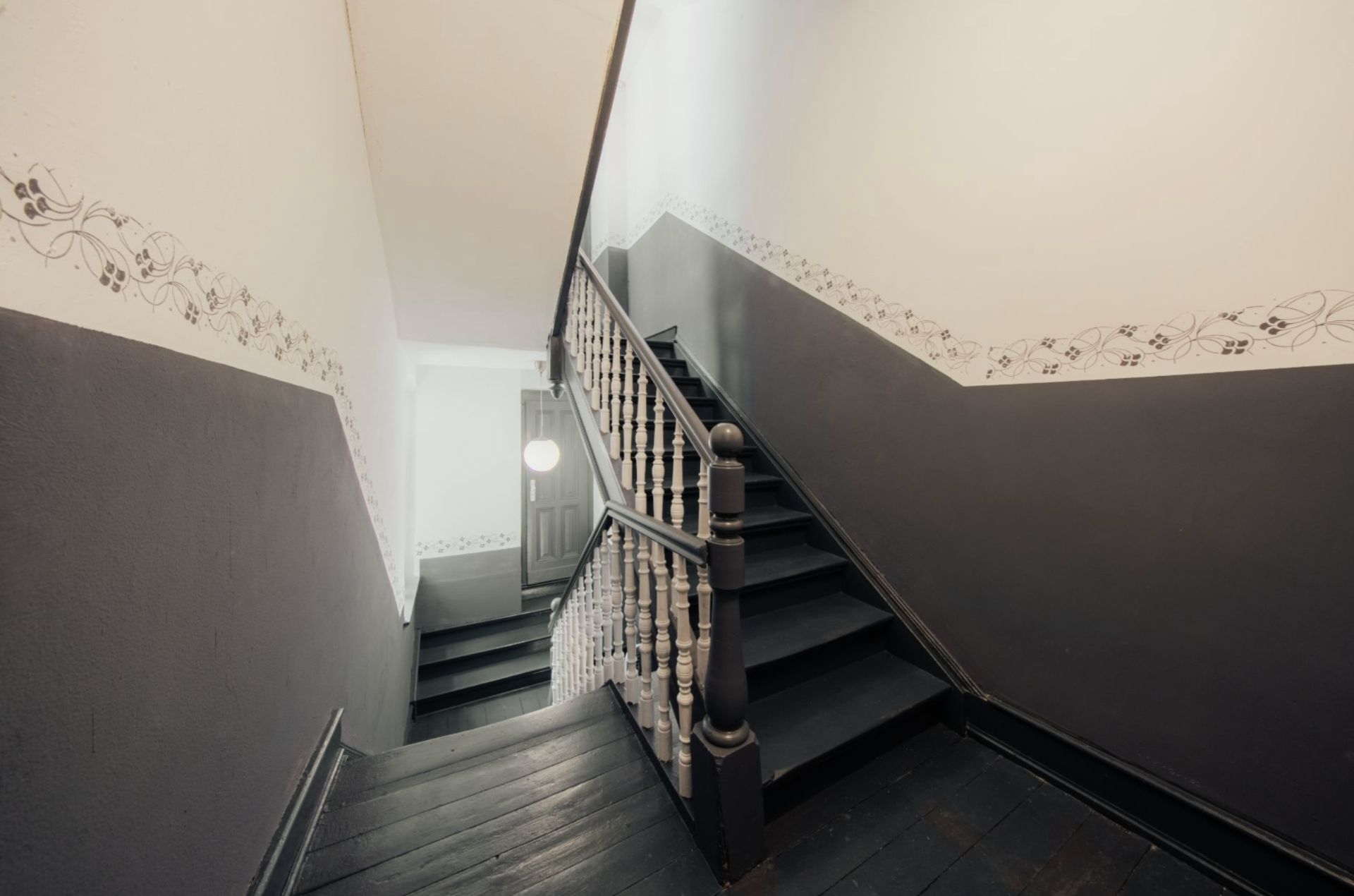 Holztreppe mit dunklem Geländer und gemusterten Wänden in einem Wohnhausflur.