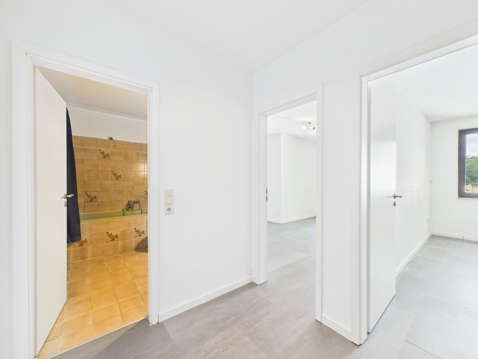 Flur mit drei offenen Türen zu Badezimmer, Wohnraum und weiterem Zimmer mit Fenster.