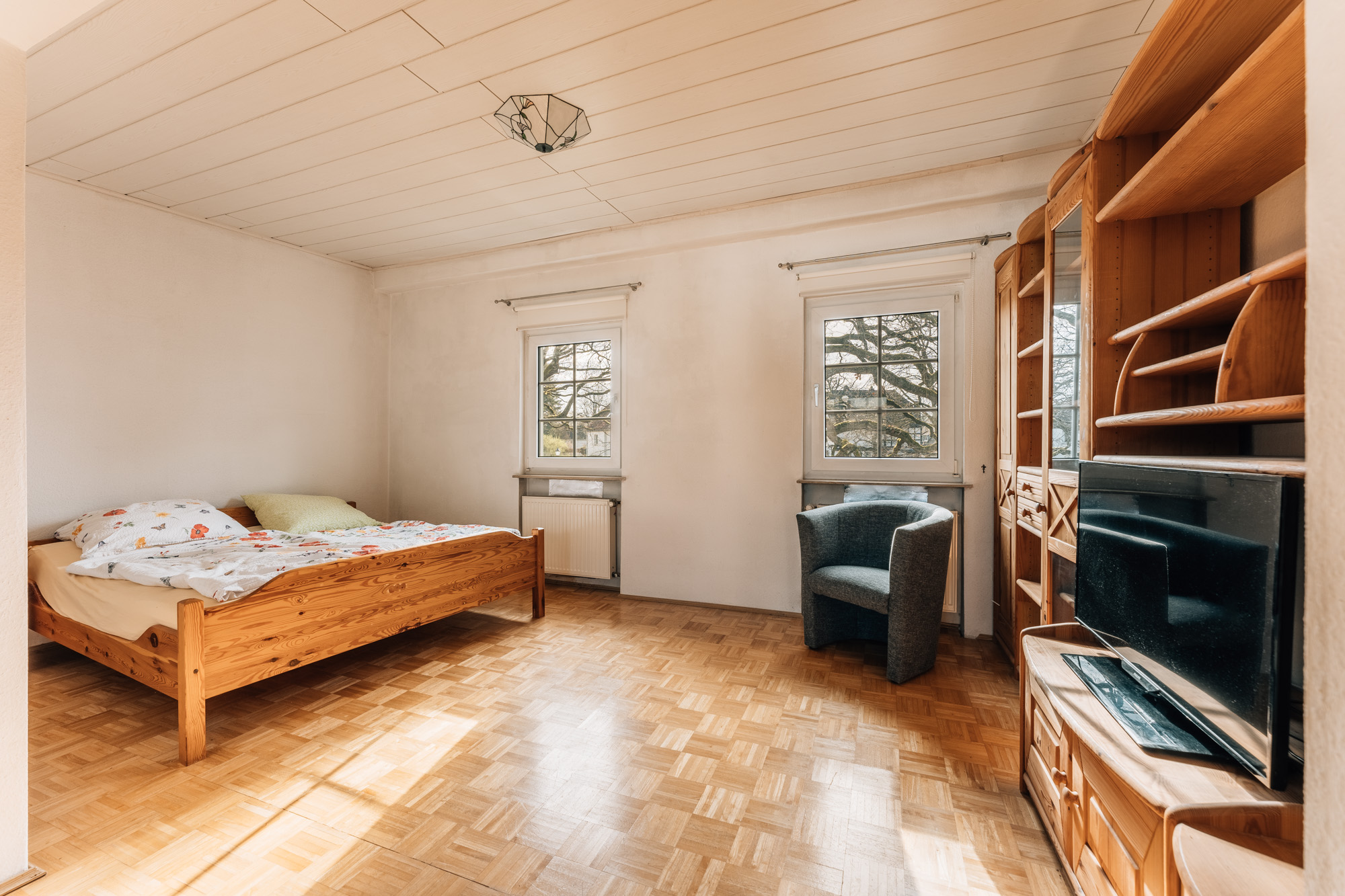Helles Schlafzimmer mit Holzbett, Sessel, Holzregal und Fernseher auf Parkettboden.