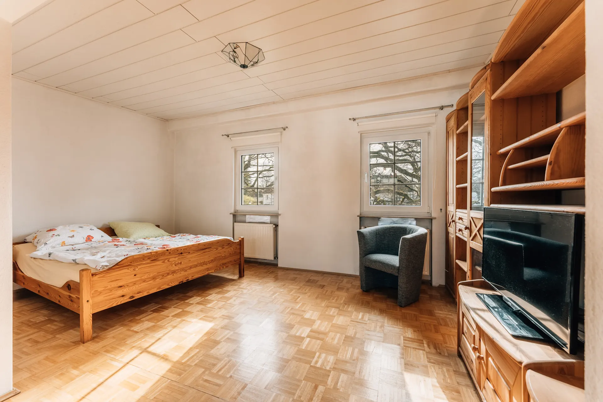 Helles Schlafzimmer mit Holzbett, Sessel, Holzregal und Fernseher auf Parkettboden.