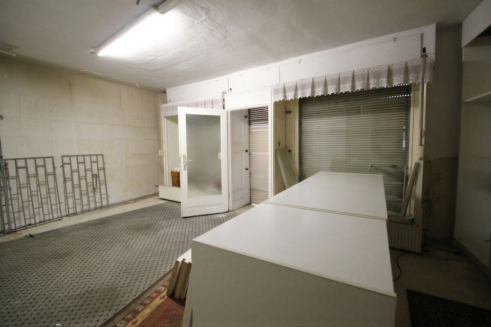 Heller Raum mit weißen Möbeln, großem Fenster mit Jalousie und offen stehender Tür.