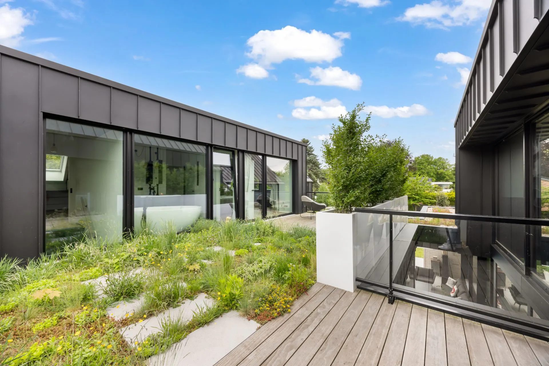 Moderne Terrasse mit Holzboden, Glasgeländer und Blick auf bepflanztes Flachdach.