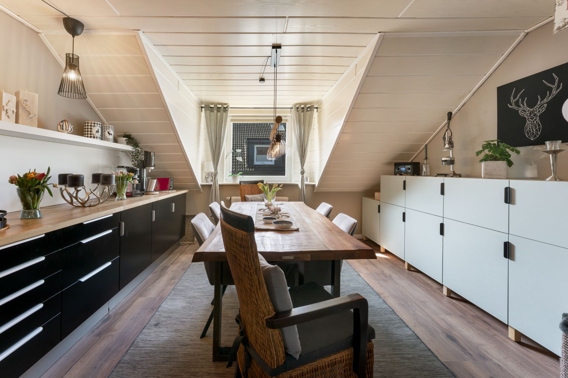 Esszimmer mit langem Holztisch, Stühlen, Kommoden, Dekoration und Fenster mit Vorhängen.