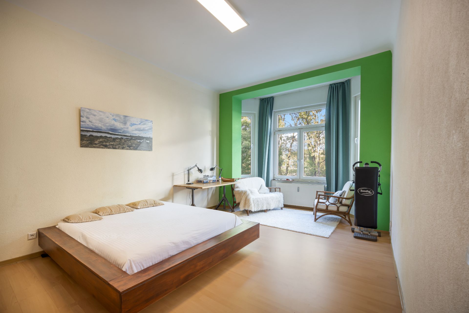 Helles Schlafzimmer mit Doppelbett, Schreibtisch, Sessel und großem Fenster mit grünen Vorhängen.