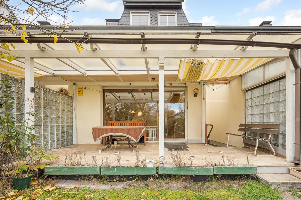 Überdachte Terrasse mit Tisch, Bank und Stühlen, angrenzend an ein Wohnhaus.