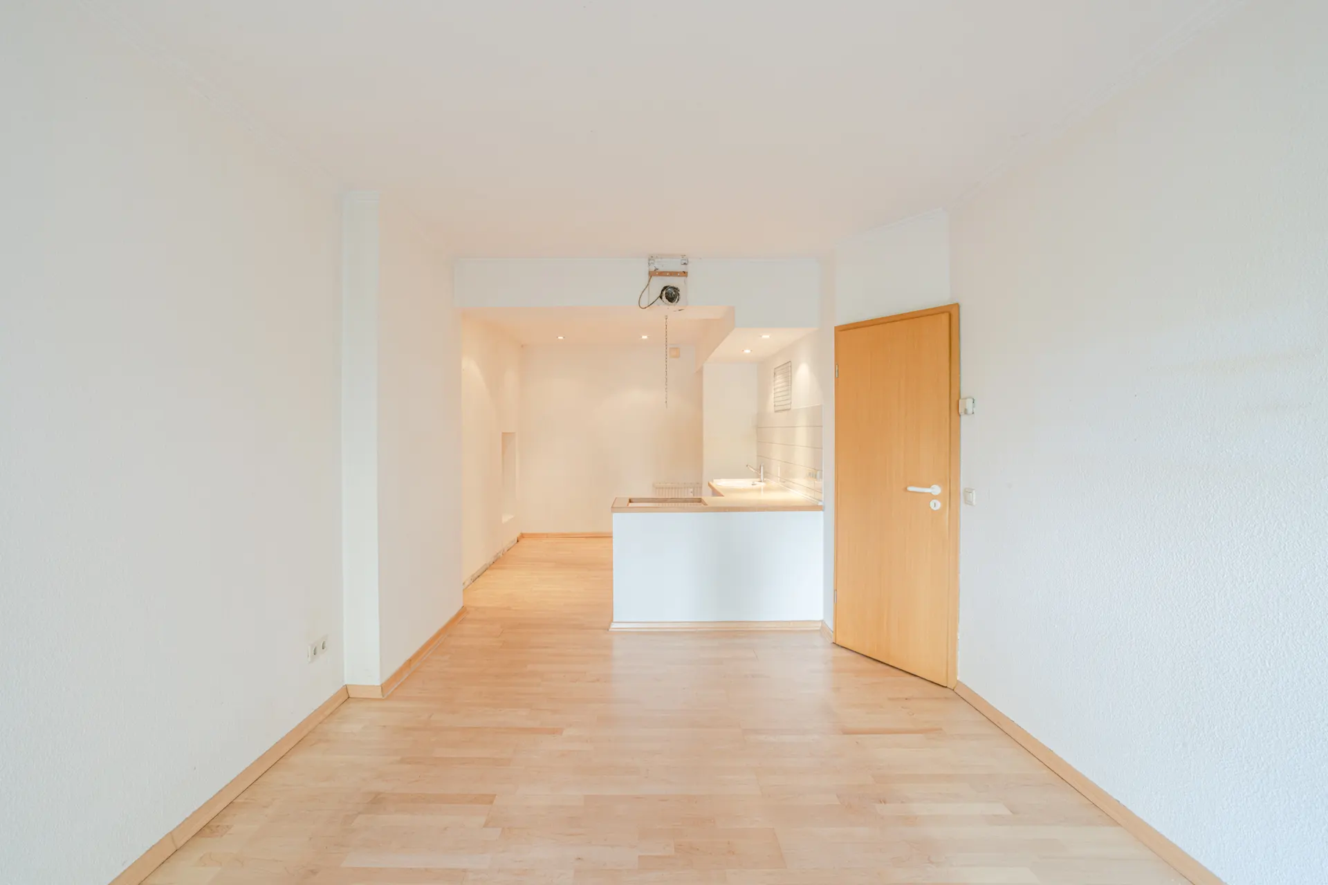 Leerer Raum mit Holzboden, offener Küchenecke und Holztür rechts.