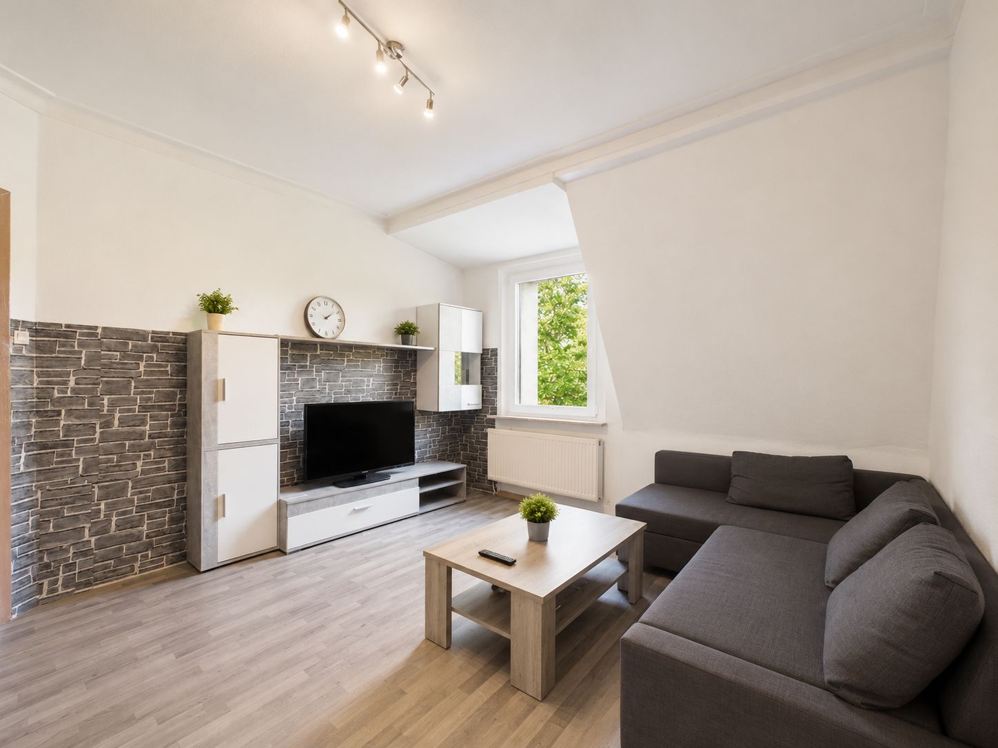 Modernes Wohnzimmer mit grauem Ecksofa, TV, hellen Möbeln und großem Fenster.