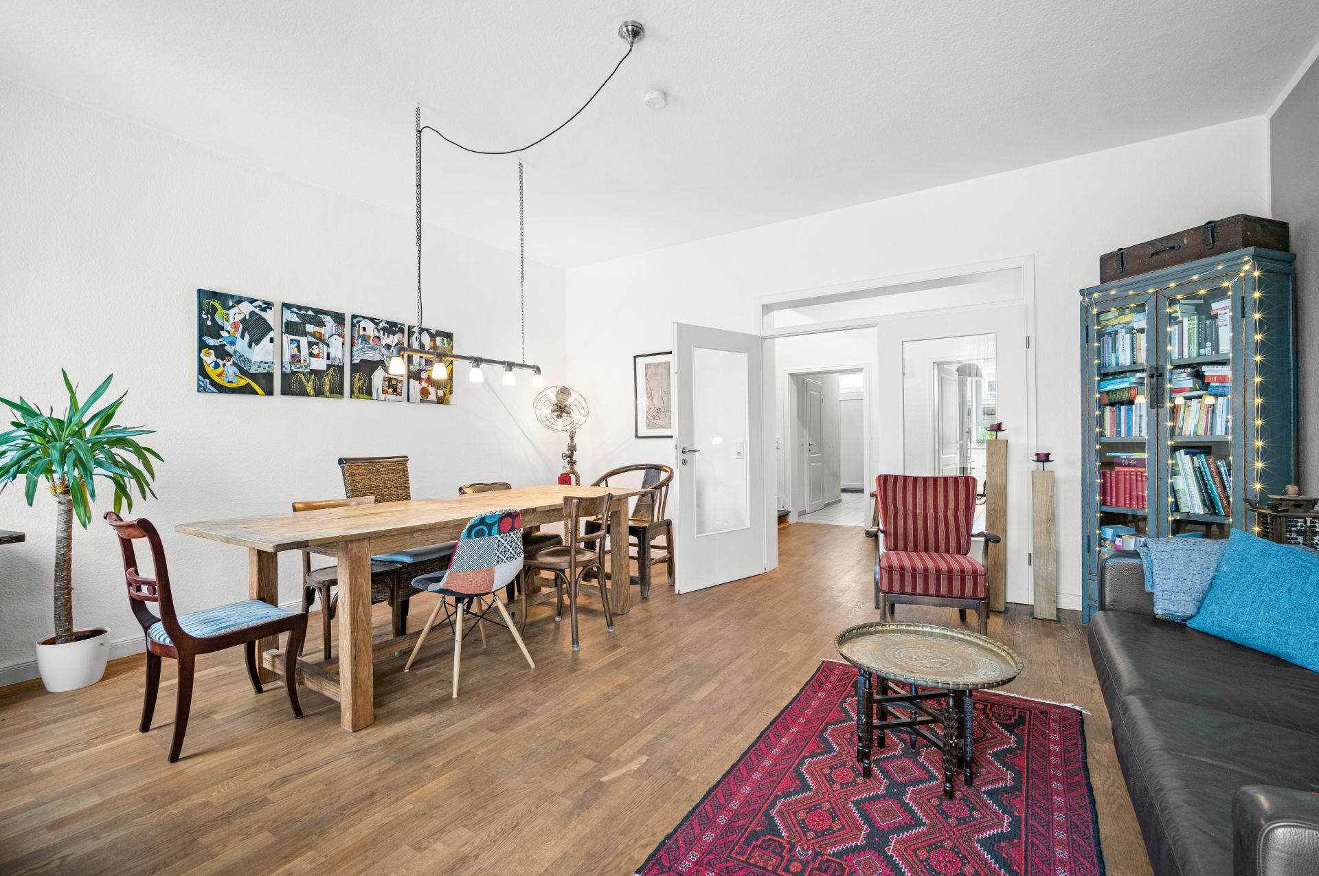 Helles Wohnzimmer mit Holzboden, Esstisch, verschiedenen Stühlen, Sofa, Teppich und Bücherregal.