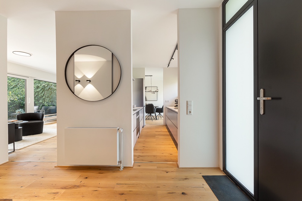 Moderner Flur mit Holzboden, Spiegel, Glastür und Blick ins Wohnzimmer.