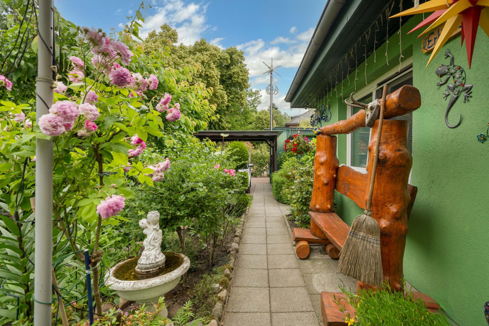 Gepflasterter Weg im Garten mit Blumen, Bank aus Holz, Brunnenfigur und grünem Haus.