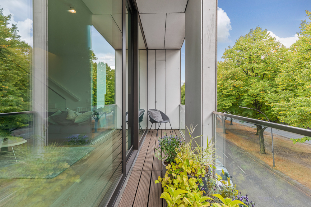 Moderner Balkon mit Holzfliesen, Pflanzen und Blick auf Bäume, zwei Stühle am Ende.