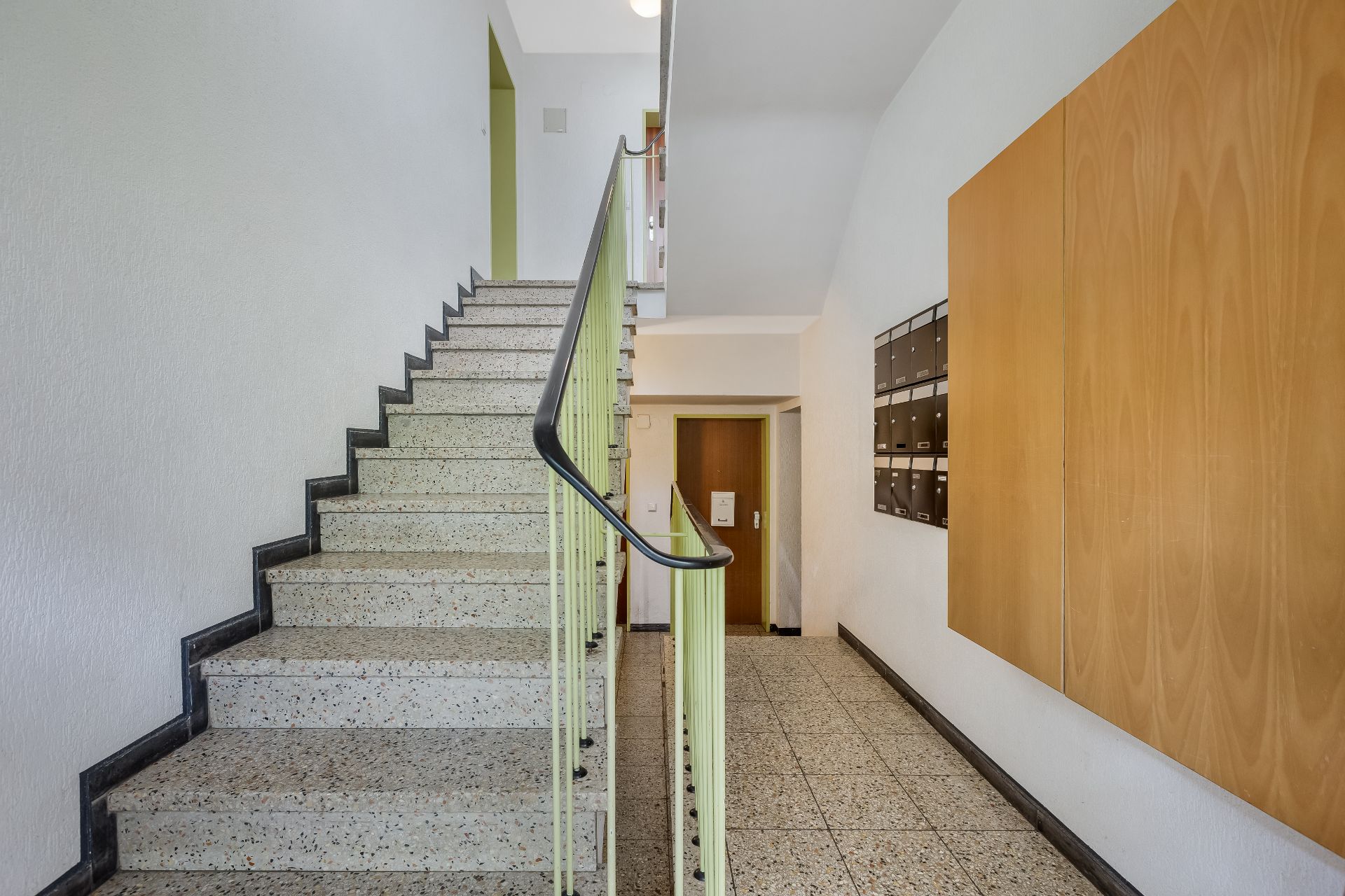 Heller Hausflur mit Treppenhaus, Briefkästen an der Wand und einer Holztür.