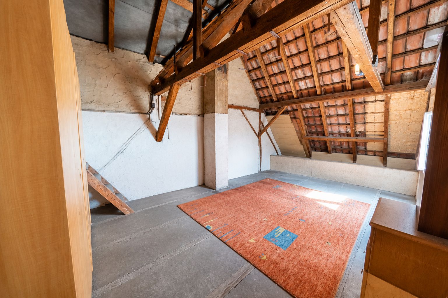 Dachzimmer mit Holzbalken, Teppich und Dachschrägen, Wände teilweise verputzt.