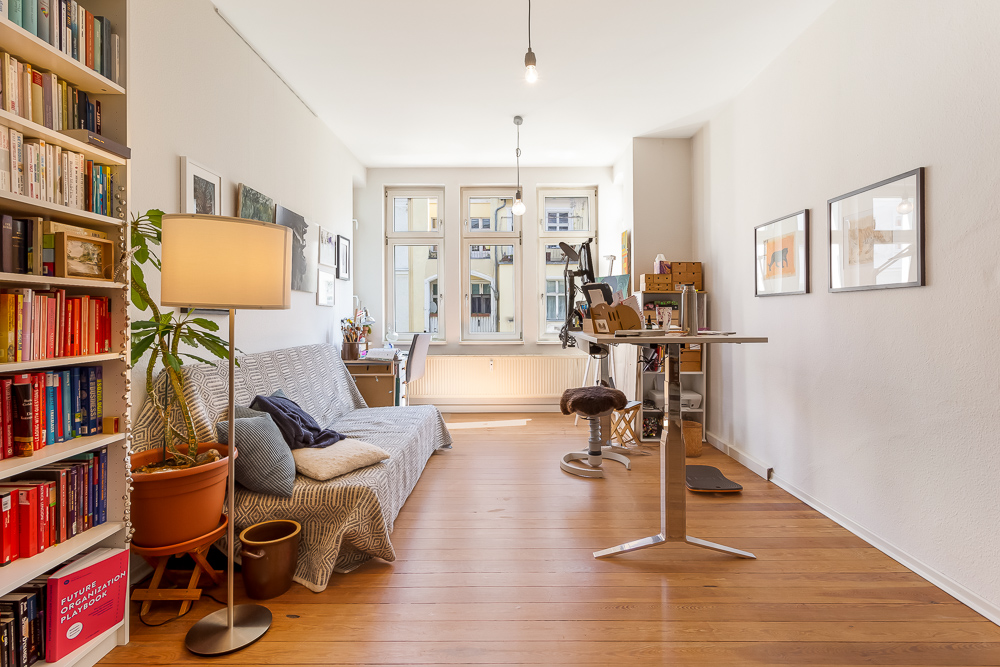Helles Wohnzimmer mit Holzboden, Sofa, Bücherregal, Schreibtisch und großen Fenstern.