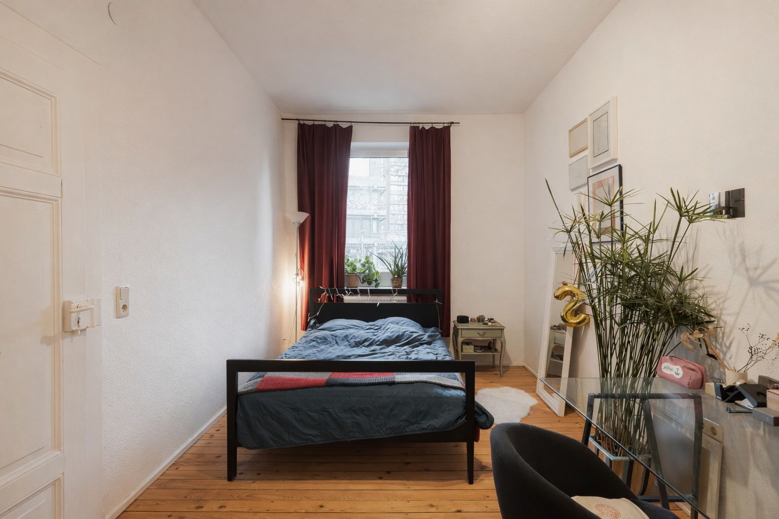 Helles Schlafzimmer mit Holzboden, Bett, rotem Vorhang und Pflanzen am Fenster.