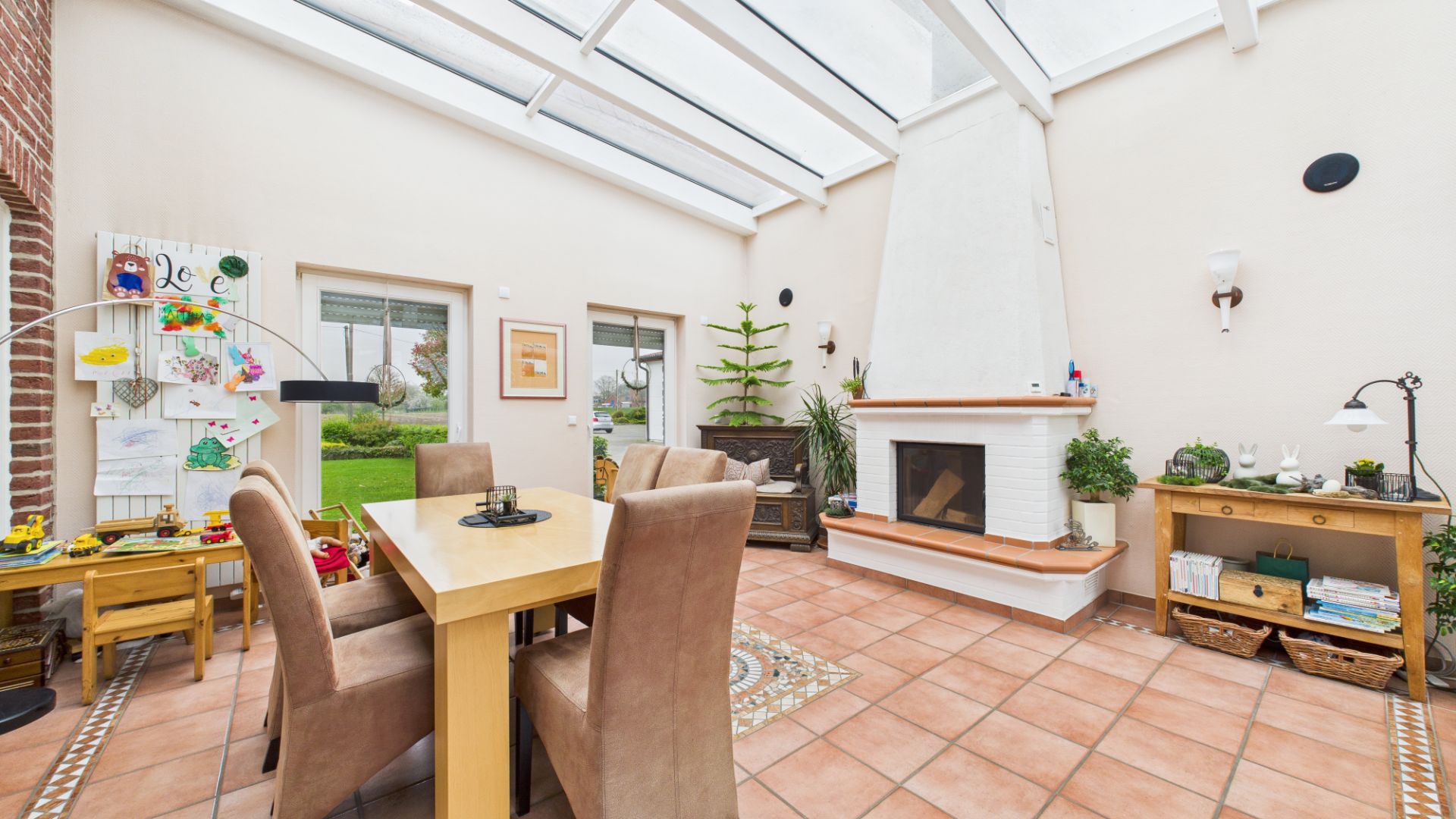 Helles Wohnzimmer mit Esstisch, Stühlen, Kamin und großen Fenstern, Spielzeug in der Ecke.