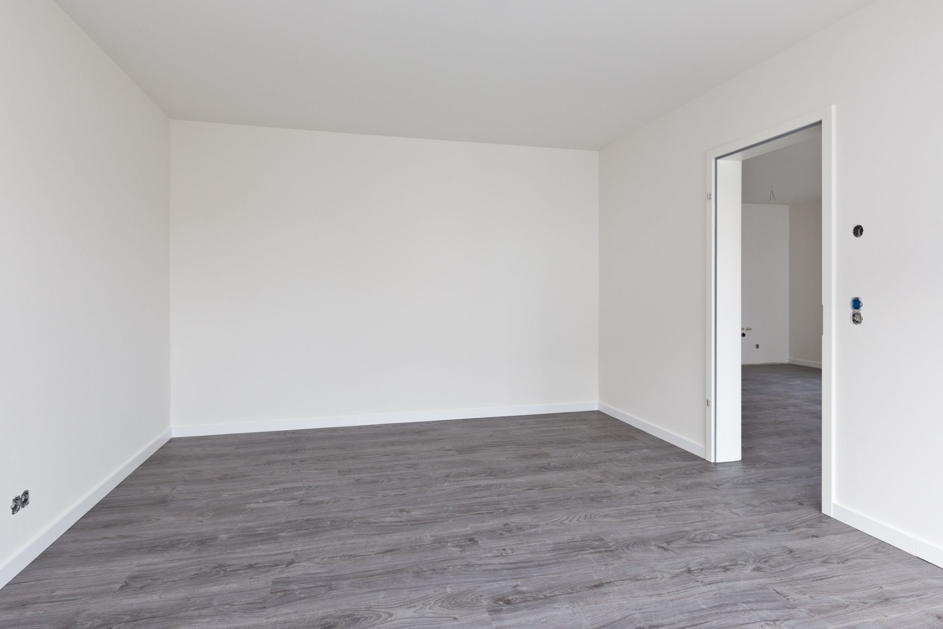 Leerer Raum mit weißen Wänden und grauem Boden, offene Tür zu weiterem Zimmer.