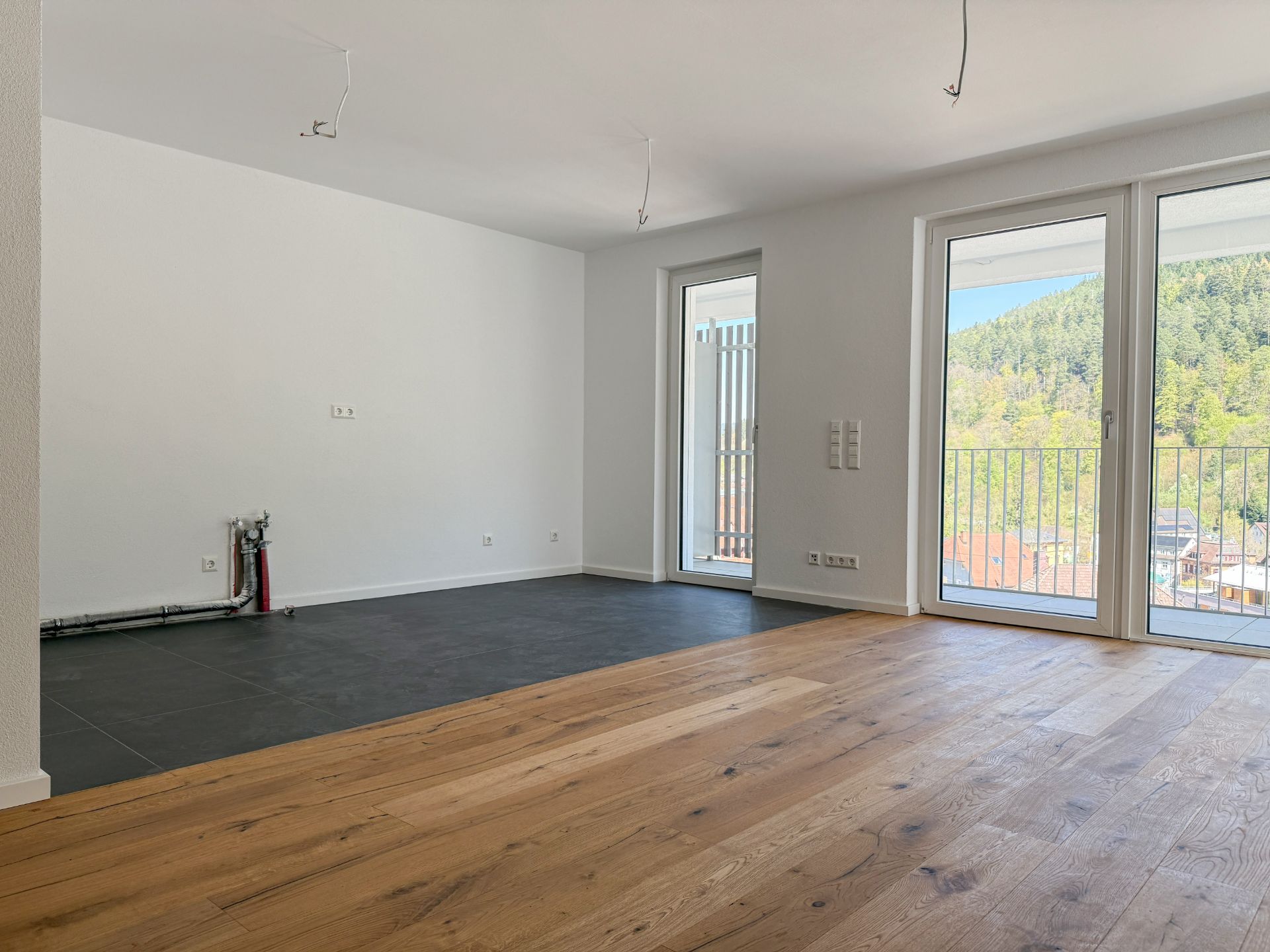 Helles Wohnzimmer mit Holzboden, bodentiefen Fenstern und Fliesenbereich.