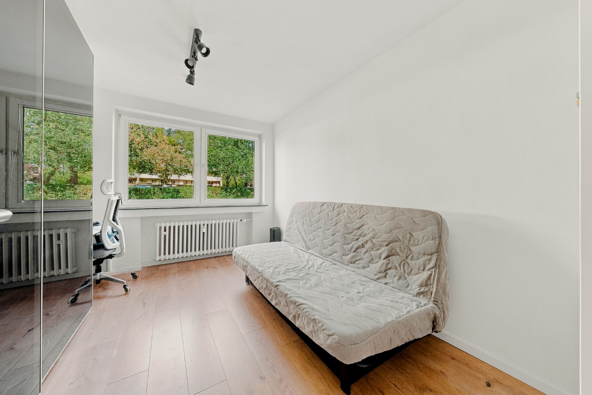 Helles Zimmer mit Schlafsofa, Schreibtisch am Fenster und Holzfußboden.