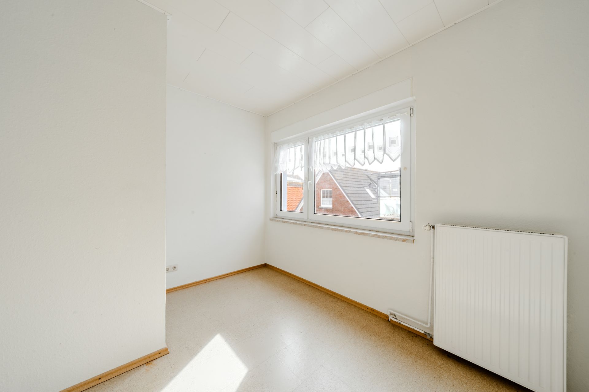 Helles, leeres Zimmer mit großem Fenster und Heizkörper an der Wand.