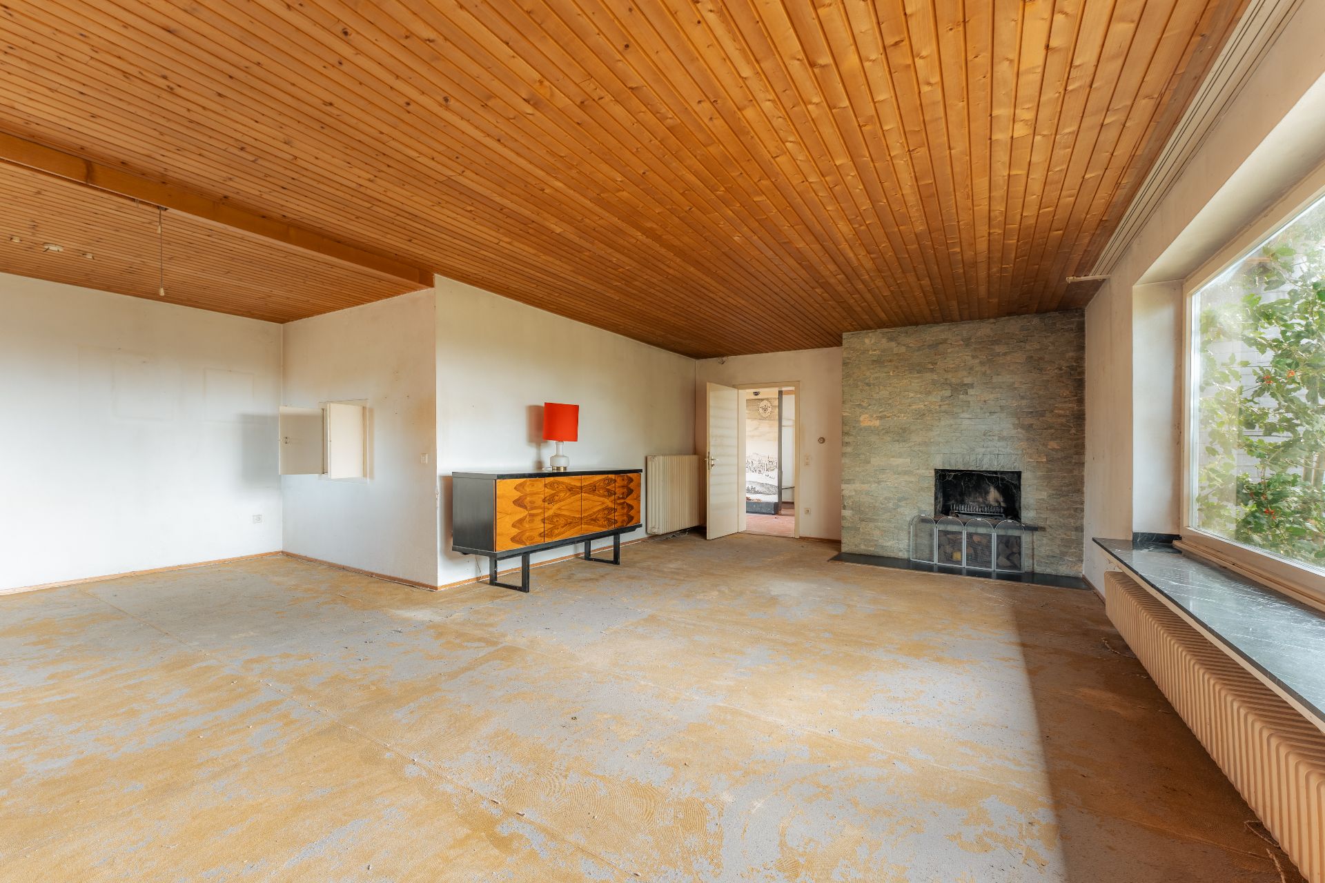 Leeres Wohnzimmer mit Holzbalkendecke, Kamin und großem Fenster.