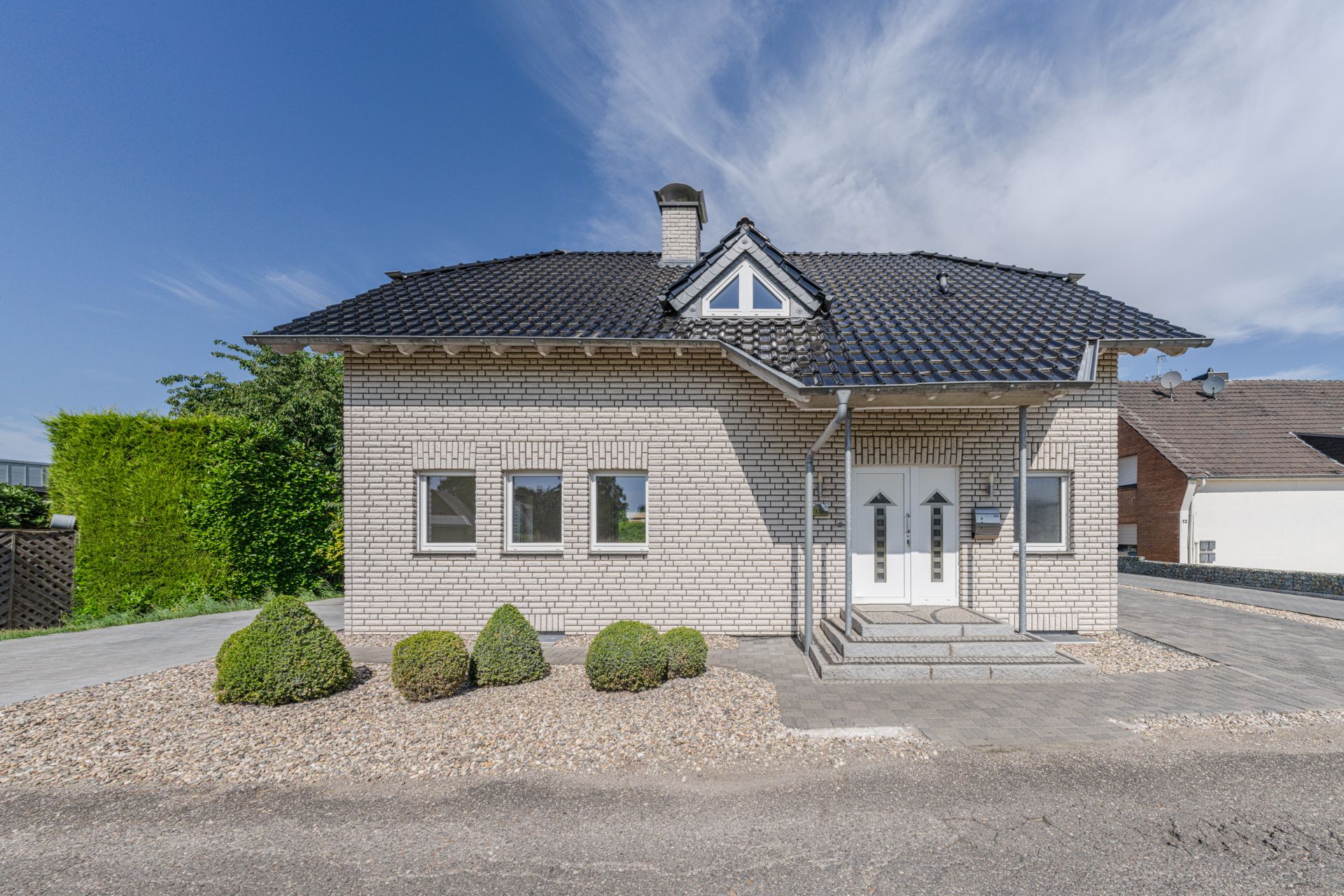 Helles Einfamilienhaus mit weißer Klinkerfassade, dunklem Dach und gepflegtem Vorgarten.