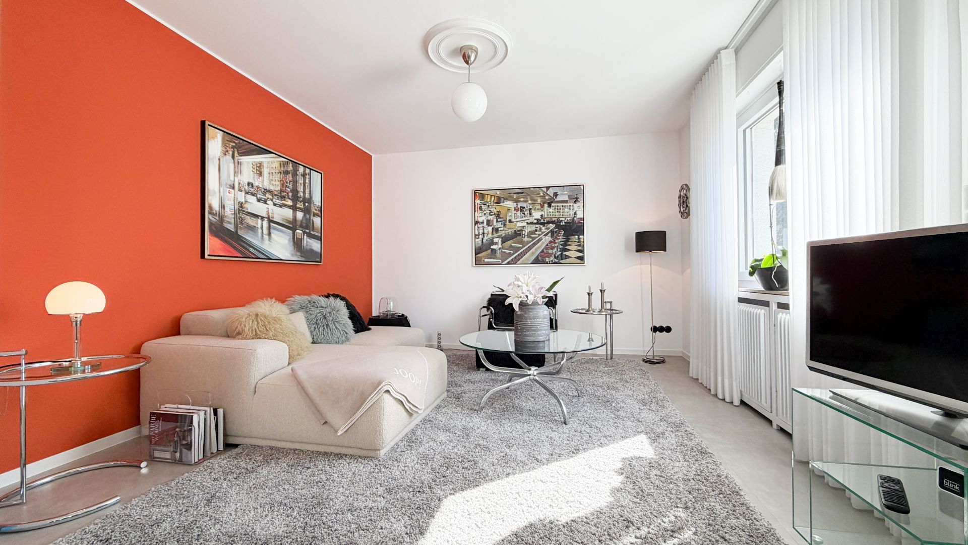 Helles Wohnzimmer mit roter Wand, Sofa, Couchtisch, Teppich und Fernseher am Fenster.