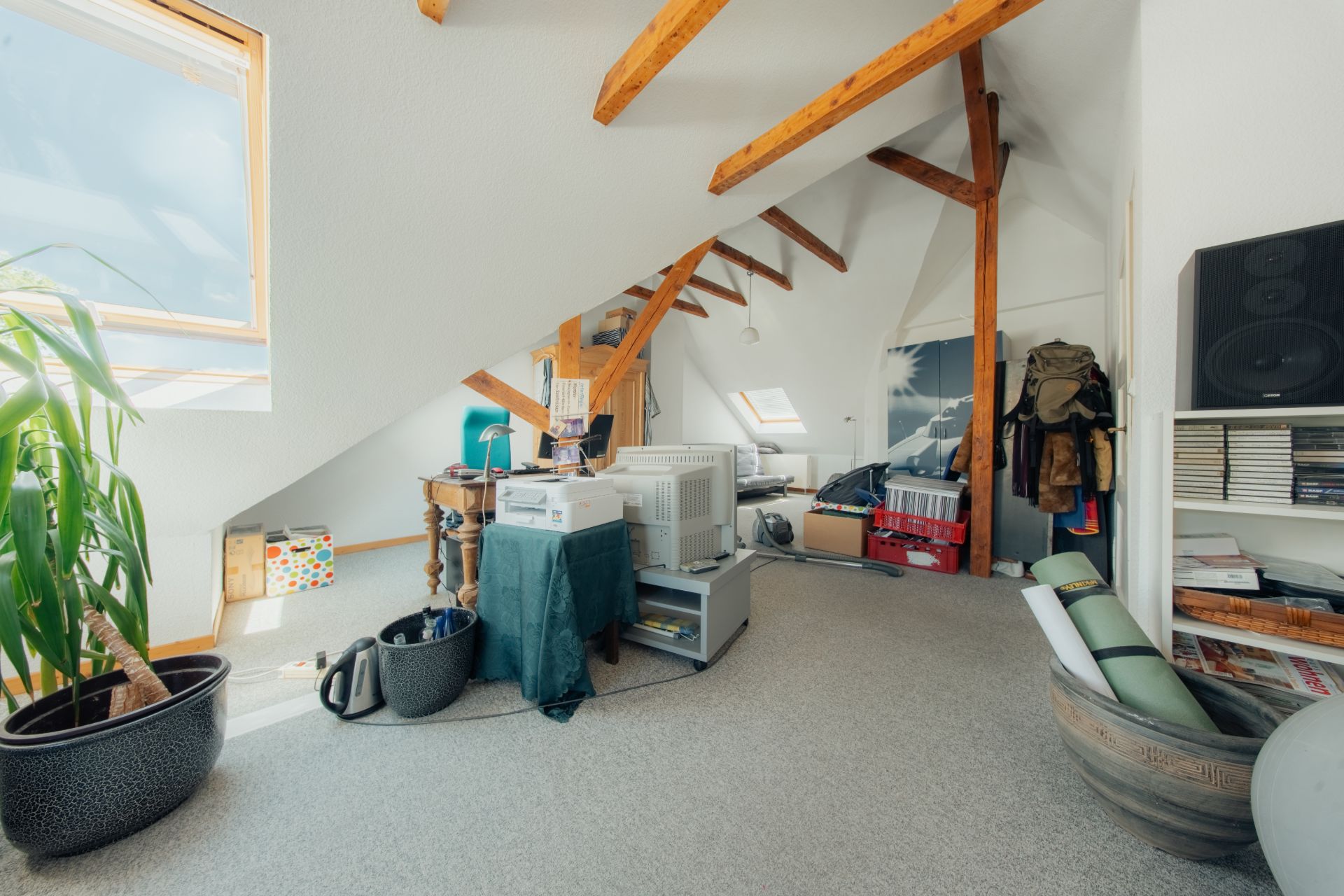 Helles Dachzimmer mit Schreibtisch, Holzbalken, Pflanzen und Regalen.