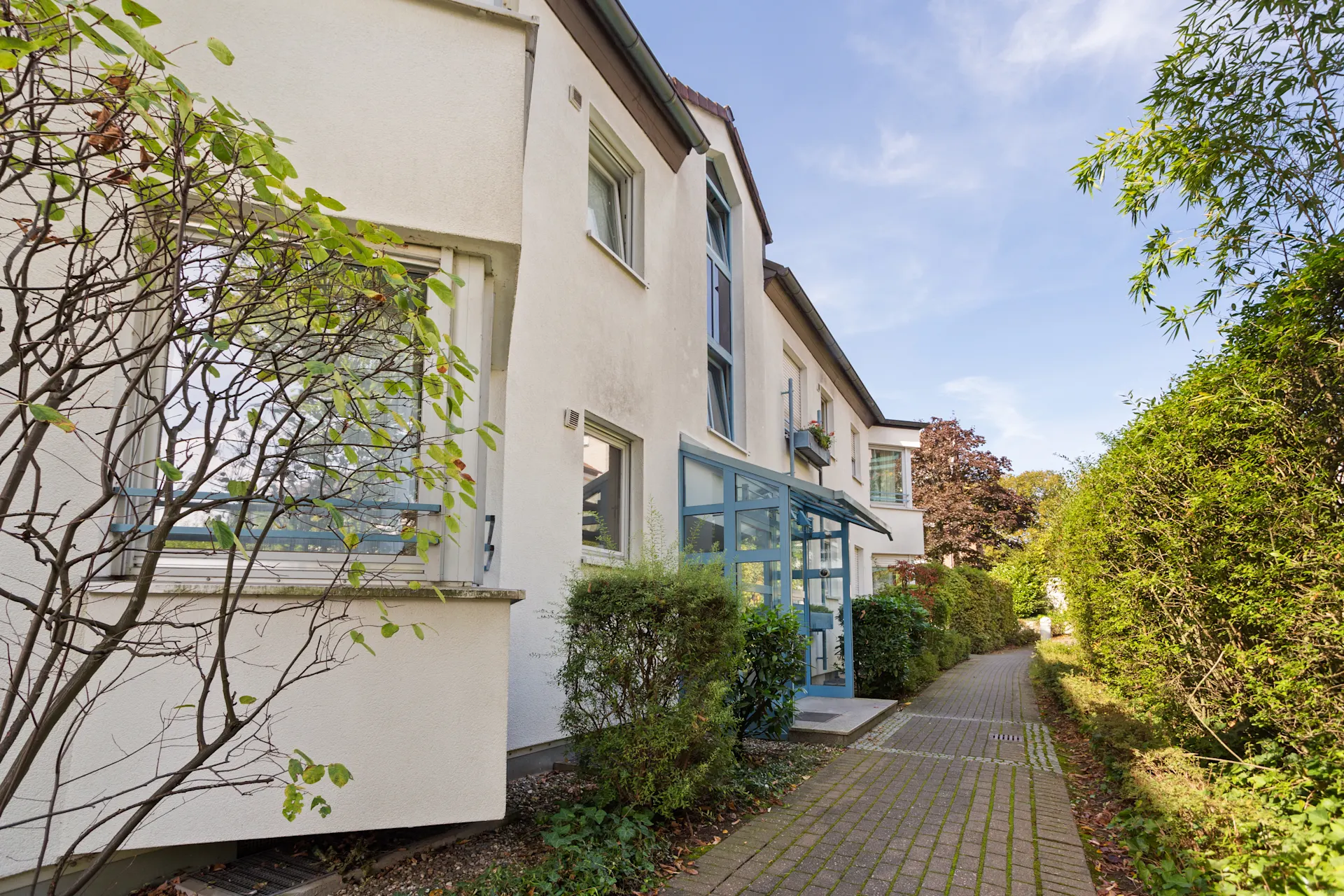 Eingangsbereich eines weißen Wohnhauses mit blauem Glasvorbau und Gartenweg.