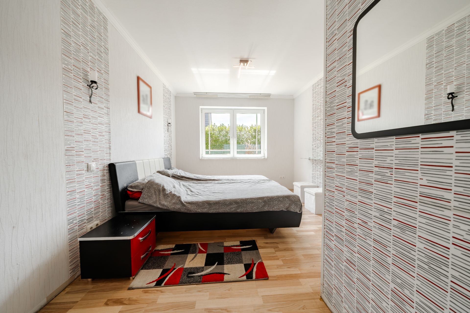 Modernes Schlafzimmer mit Doppelbett, Nachttisch, Teppich und großem Fenster, helle Wände und Holzfußboden.