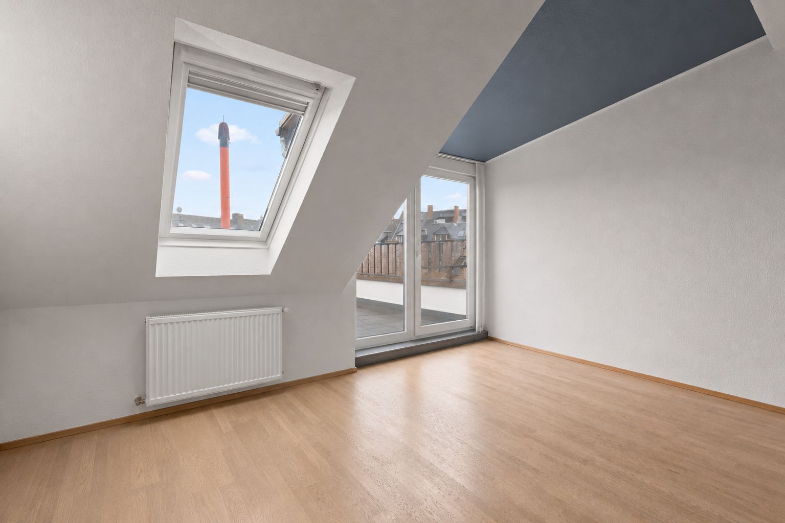 Helles Dachzimmer mit schrägem Fenster, Holzboden und Zugang zu einem Balkon.