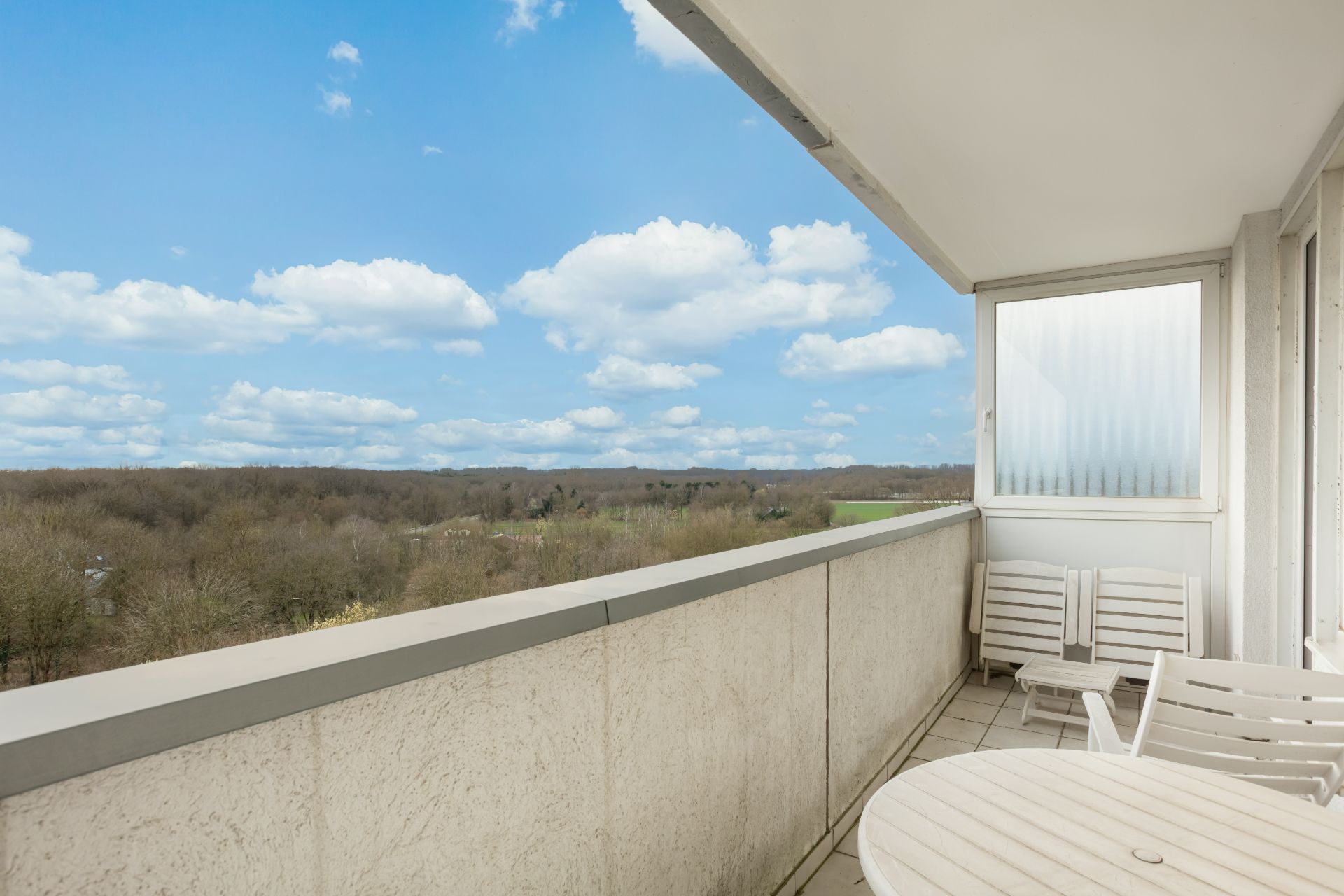 Balkon mit weißen Gartenmöbeln und Ausblick auf Wald und Wiese bei blauem Himmel.