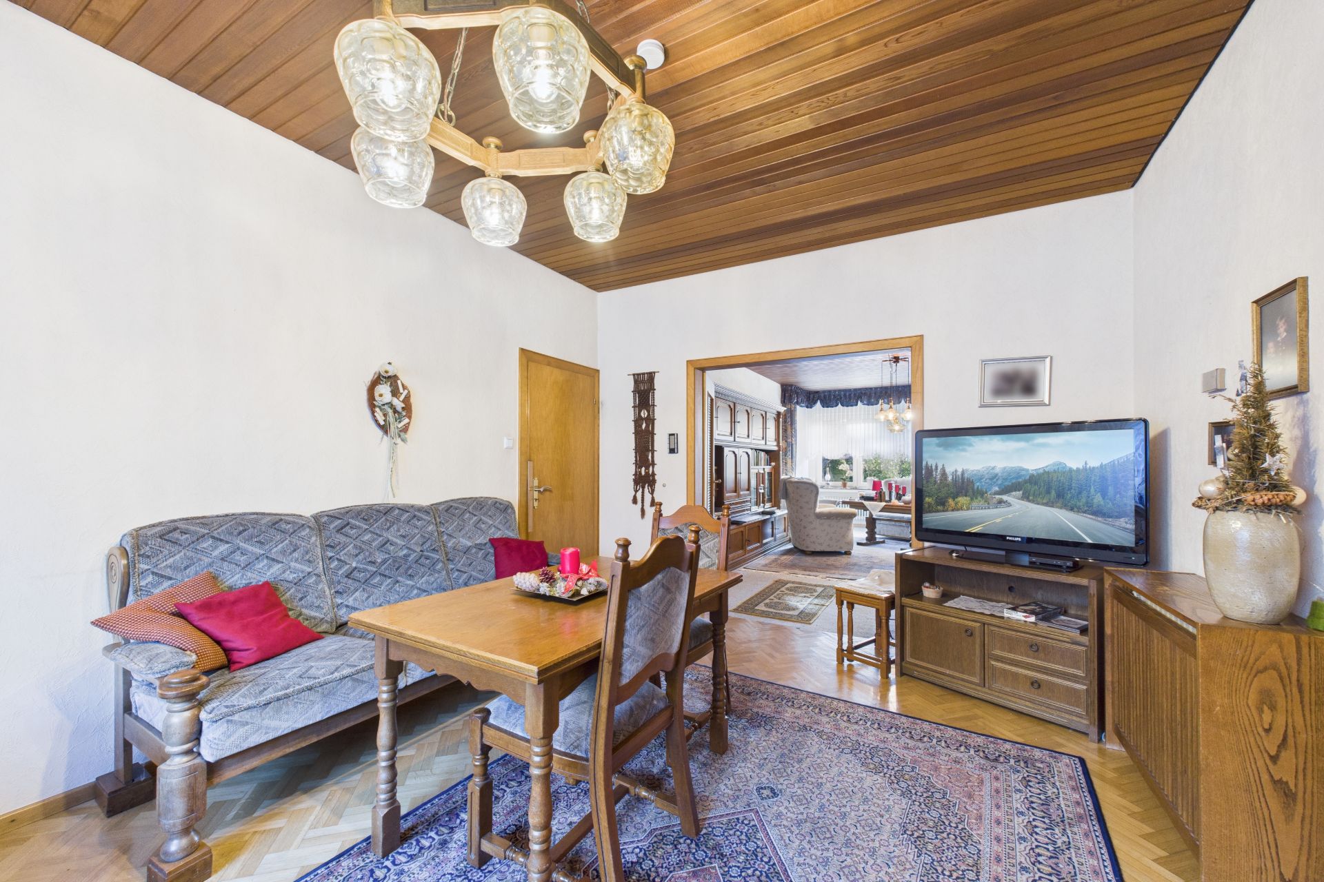 Wohnzimmer mit Holztisch, Stühlen, Sofa, Fernseher und Teppich, angrenzender Raum sichtbar.